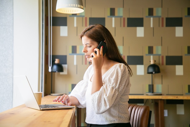 Libero professionista femminile concentrato che lavora al computer portatile e che comunica sul telefono delle cellule nello spazio di co-working