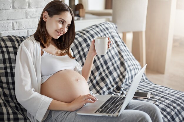 Libero professionista in gravidanza felice che utilizza un laptop per eseguire le ultime modifiche al design. Lavorare da remoto.