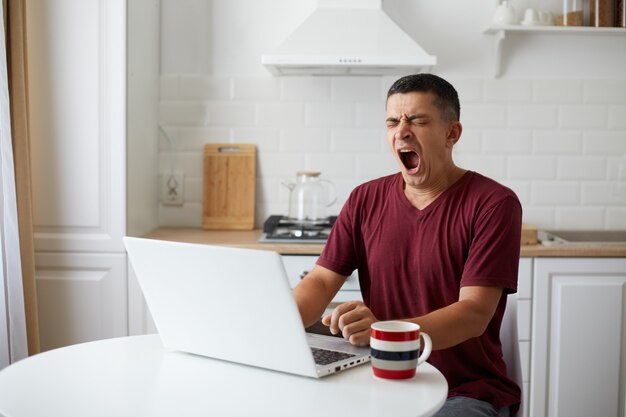 Libero professionista stanco seduto al tavolo in cucina, avendo una notte insonne lavorando online, sbadigliando, si sente assonnato, guardando il taccuino, bevendo caffè per ottenere energia.