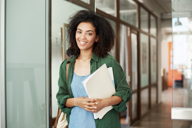 Libri sorridenti della tenuta della bella studentessa africana allegra in università. Concetto di educazione.