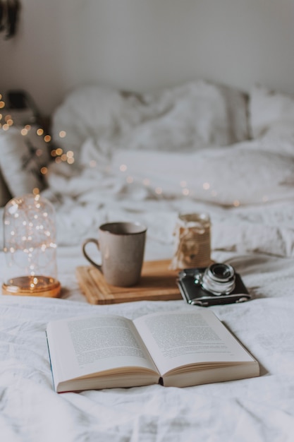 Libro aperto, una macchina fotografica, un vassoio e una tazza su un letto con lenzuola bianche