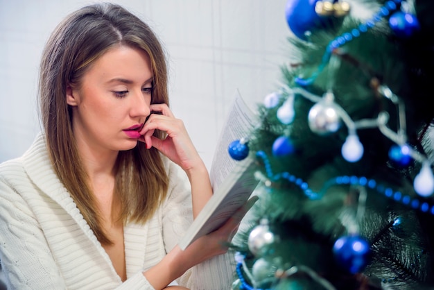 Libro di lettura della donna nella casa decorata di natale. Concetto di festa, vacanze, tempo libero, letteratura e concetto di persone