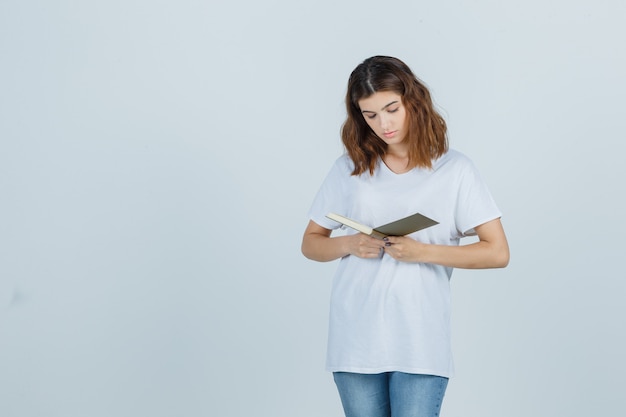 Libro di lettura della giovane signora mentre levandosi in piedi in maglietta, jeans e guardando concentrato. vista frontale.