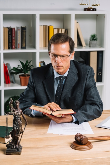 Libro di lettura maschio maturo dell'avvocato con la statua della giustizia e del martelletto sulla tavola di legno