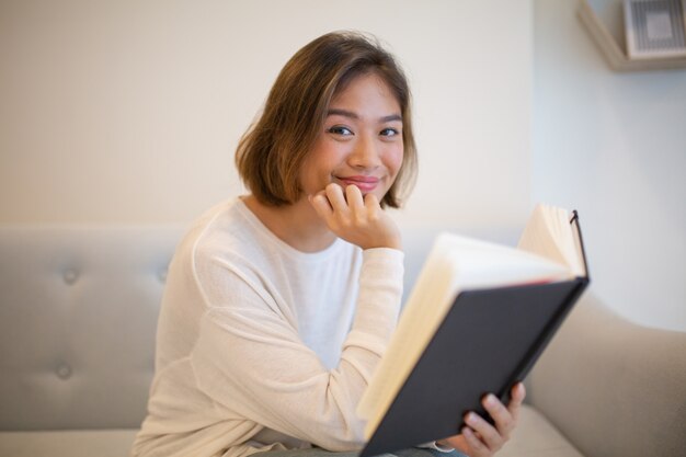 Libro di lettura sorridente della giovane donna sul sofà a casa