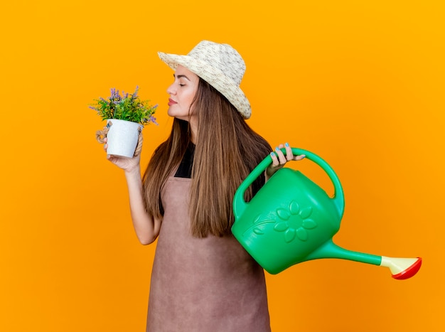 Lieta bella ragazza giardiniere che indossa uniforme e cappello da giardinaggio tenendo annaffiatoio e annusando fiore in vaso di fiori in mano isolato su sfondo arancione