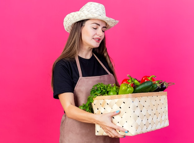 Lieta bella ragazza giardiniere in uniforme che indossa cappello da giardinaggio tenendo e guardando il cesto di verdure isolato su sfondo rosa