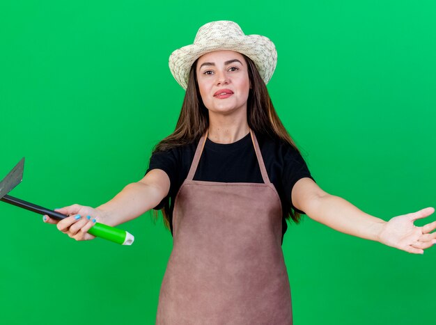 Lieta bella ragazza giardiniere in uniforme che indossa cappello da giardinaggio tenendo la zappa rastrello e tendendo le mani isolate su verde