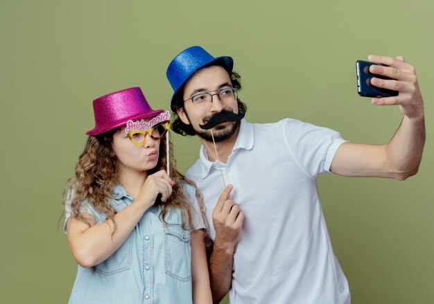 Lieta coppia giovane indossando cappello rosa e blu prendere una ragazza selfie con maschera per gli occhi mascherata su stick e guy holding baffi finti su stick