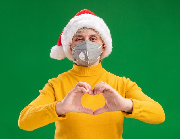 Lieta donna anziana con santa hat indossando maschera medica gesticolando segno di cuore isolato su sfondo verde con copia spazio