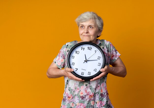 Lieta donna anziana tiene l'orologio guardando il lato isolato sulla parete arancione