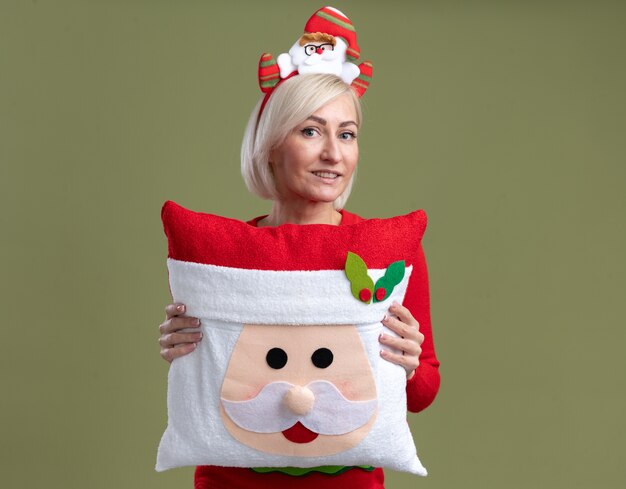 Lieta donna bionda di mezza età che indossa la fascia di Babbo Natale e maglione di Natale che tiene il cuscino di Babbo Natale guardando la telecamera guardando isolato su sfondo verde oliva con spazio di copia