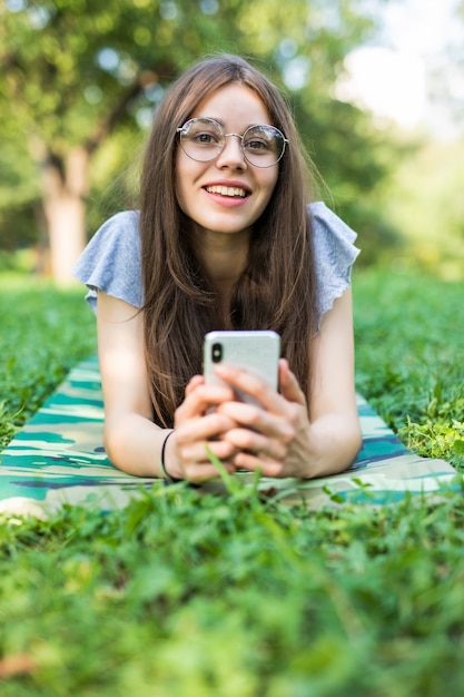 Lieta donna bruna in occhiali sdraiato sull'erba e utilizza lo smartphone nel parco