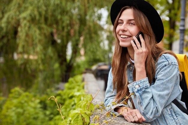 Lieta donna dai capelli scuri parla al cellulare nel parco cittadino, posa sopra albero verde sfocato, gode di una bella chiacchierata, indossa cappello e giacca di jeans