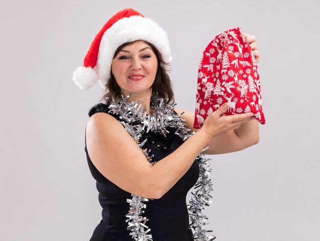 Lieta donna di mezza età che indossa santa hat e orpello ghirlanda intorno al collo tenendo il regalo di Natale sacco guardando la telecamera isolata su sfondo bianco