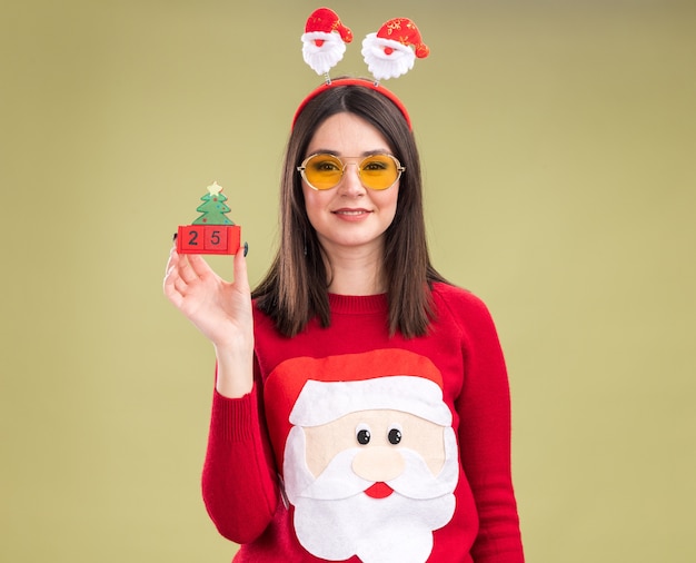 Lieta giovane bella ragazza caucasica che indossa il maglione di Babbo Natale e la fascia con gli occhiali che tiene il giocattolo dell'albero di Natale con la data che guarda l'obbiettivo isolato su fondo verde oliva