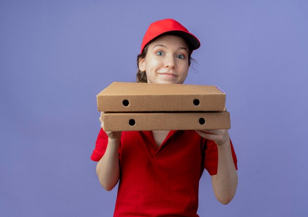 Lieta giovane bella ragazza delle consegne che indossa l'uniforme rossa e il cappuccio che tiene i pacchetti di pizza guardando la telecamera isolata su sfondo viola con spazio di copia