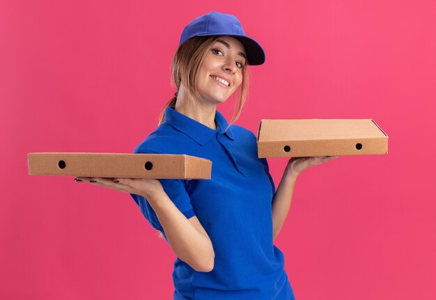 Lieta giovane bella ragazza delle consegne in uniforme tiene scatole per pizza su due mani sul rosa