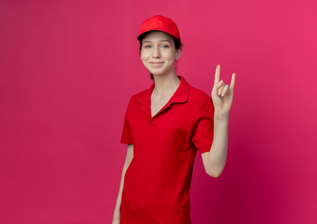 Lieta giovane bella ragazza di consegna che indossa l'uniforme rossa e cappuccio facendo segno di roccia isolato su sfondo cremisi con copia spazio