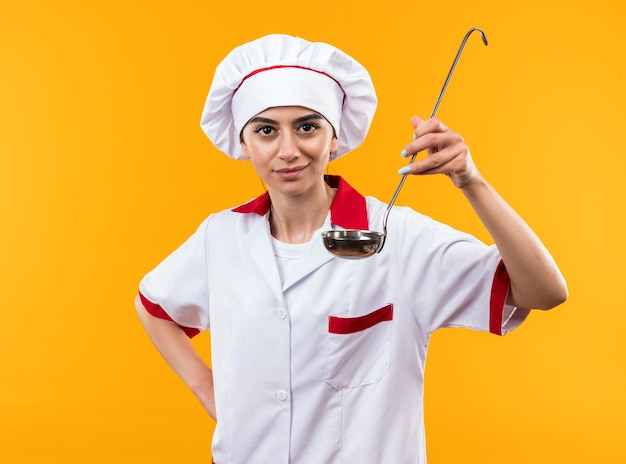 Lieta giovane bella ragazza in uniforme da chef che tiene mestolo mettendo la mano sull'anca isolata sulla parete arancione