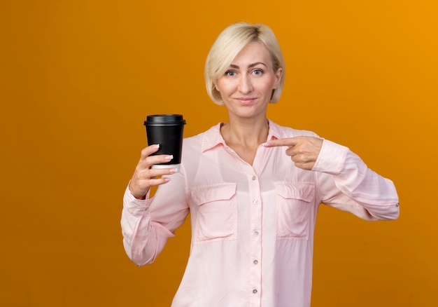 Lieta giovane bionda donna slava azienda e punti al poliziotto di caffè isolato sulla parete arancione