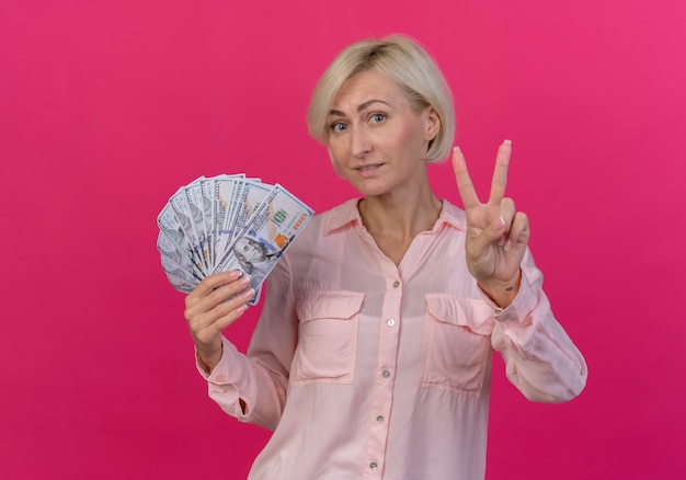 Lieta giovane bionda donna slava tenendo i soldi e facendo segno di pace isolato su sfondo rosa