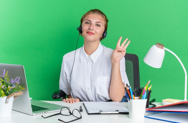 Lieta giovane bionda ragazza del call center che indossa la cuffia seduta alla scrivania con strumenti di lavoro che mostra tre con la mano