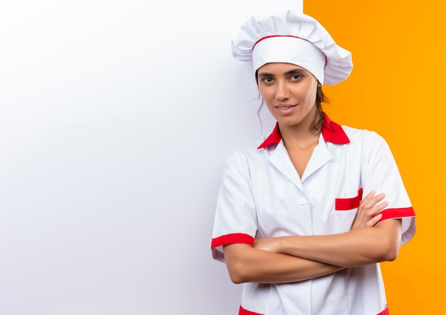 Lieta giovane cuoca che indossa l'uniforme dello chef in piedi con il muro bianco e le mani incrociate con lo spazio della copia