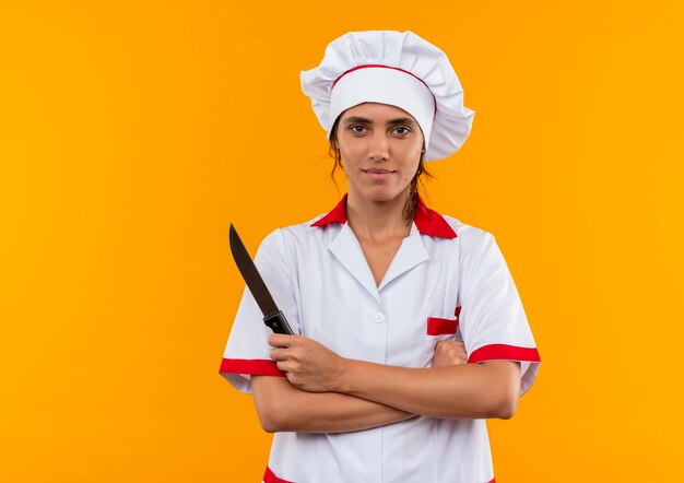 Lieta giovane cuoca che indossa uniforme da chef tenendo il coltello e incrocio le mani sulla parete gialla isolata con spazio di copia