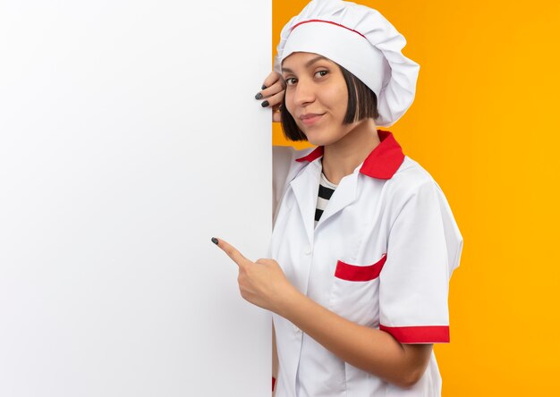 Lieta giovane cuoca in uniforme da chef in piedi dietro e indicando il muro bianco isolato sul muro