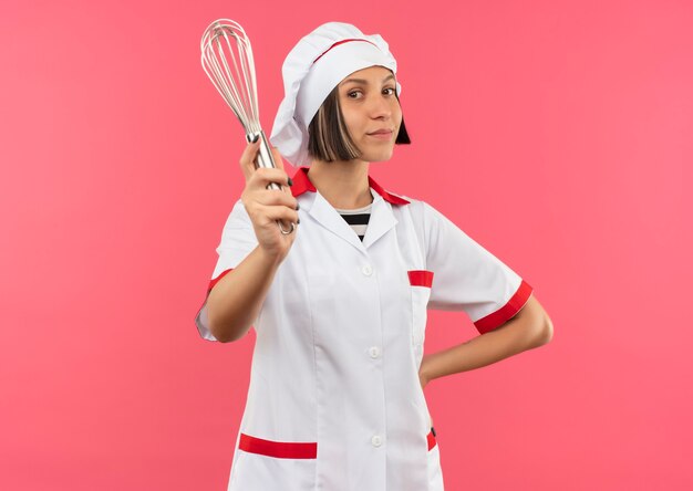Lieta giovane cuoca in uniforme da chef mettendo la mano sulla vita e allungando la frusta verso la parte anteriore isolata sul muro rosa