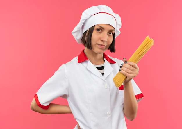 Lieta giovane cuoca in uniforme da chef tenendo gli spaghetti e tenendo la mano dietro la schiena isolata sulla parete rosa