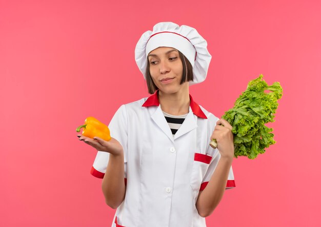 Lieta giovane cuoca in uniforme da chef tenendo pepe e lattuga e guardando pepe isolato sulla parete rosa