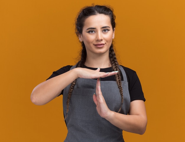 Lieta giovane donna barbiere in uniforme che mostra il gesto di timeout isolato sulla parete arancione