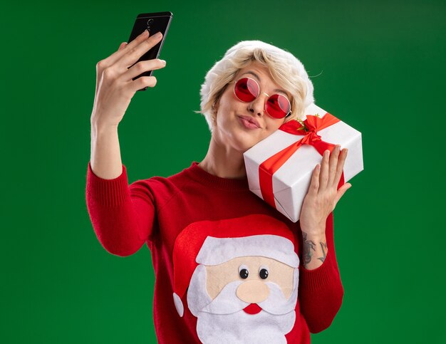Lieta giovane donna bionda che indossa un cappello di Natale e Babbo Natale maglione di Natale con gli occhiali che tiene il pacchetto regalo toccando il viso con esso prendendo selfie isolato su sfondo verde
