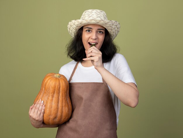 Lieta giovane donna bruna giardiniere in uniforme che indossa il cappello da giardinaggio tiene la zucca e finge di mordere il cetriolo isolato sulla parete verde oliva