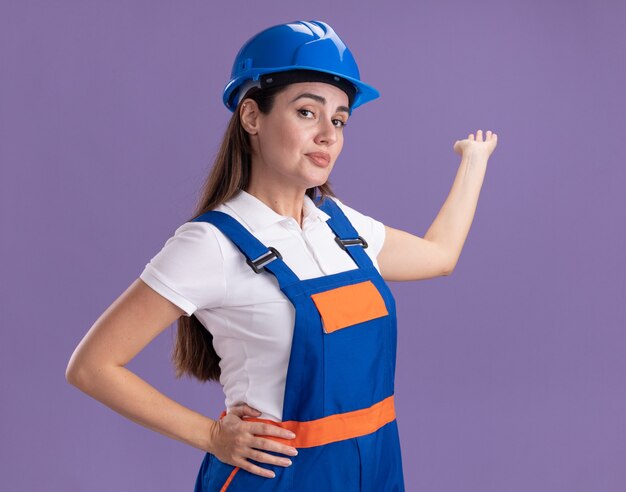 Lieta giovane donna costruttore in punti uniformi con la mano dietro isolato sulla parete viola con spazio di copia
