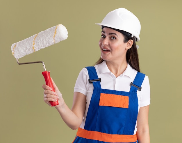 Lieta giovane donna costruttore in uniforme tenendo la spazzola a rullo isolata sulla parete verde oliva