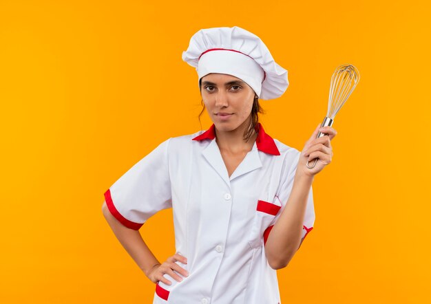 Lieta giovane donna cuoca che indossa uniforme da chef tenendo la frusta e mettendo la mano sul fianco con lo spazio della copia