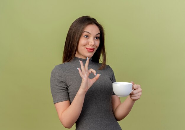 Lieta giovane donna graziosa che tiene tazza guardando dritto e facendo segno ok isolato su sfondo verde oliva con spazio di copia