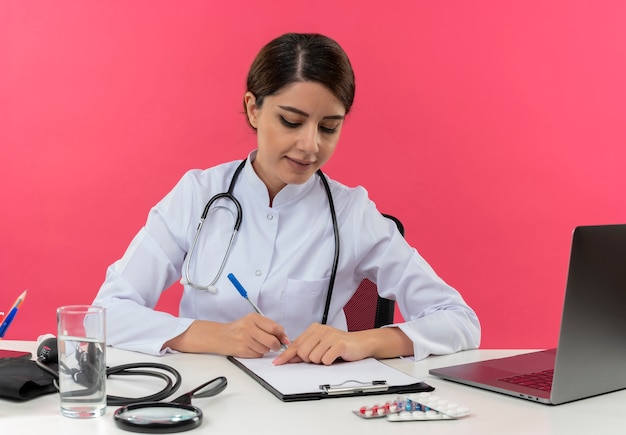 Lieta giovane donna medico indossando abito medico con uno stetoscopio seduto alla scrivania lavora sul computer con strumenti medici scrivere qualcosa negli appunti con spazio di copia