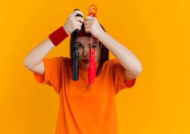 Lieta giovane donna sportiva che indossa la fascia e braccialetti tenendo le corde per saltare tenendoli vicino al viso isolato sulla parete arancione con spazio di copia