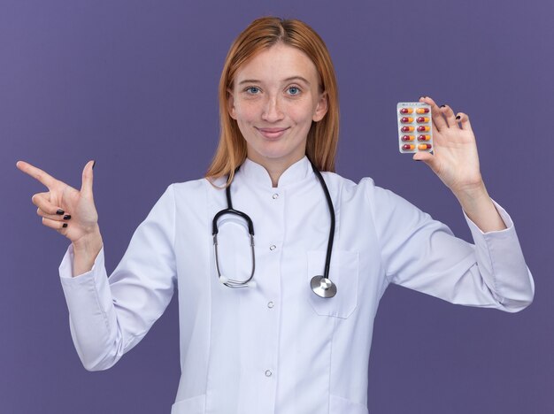 Lieta giovane dottoressa allo zenzero che indossa una tunica medica e uno stetoscopio che mostra un pacco di pillole mediche che puntano a lato isolato su parete viola