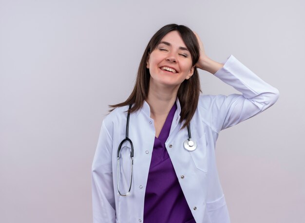 Lieta giovane dottoressa in abito medico con lo stetoscopio mette la mano dietro la testa su sfondo bianco isolato con spazio di copia