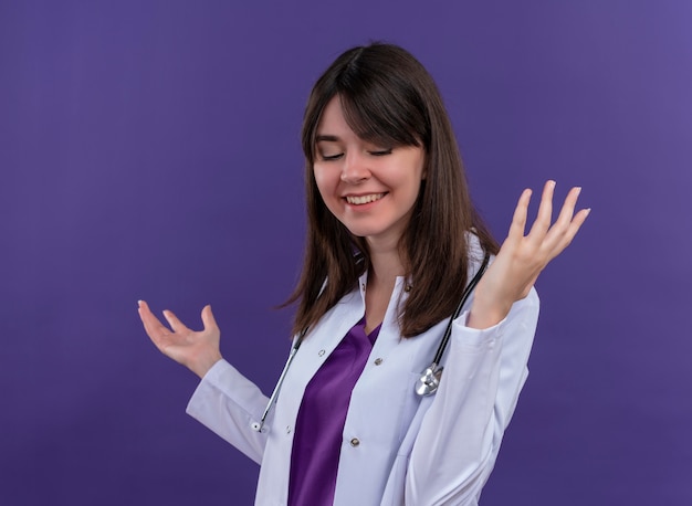 Lieta giovane dottoressa in abito medico con lo stetoscopio tiene le mani aperte su sfondo viola isolato con spazio di copia