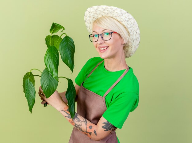Lieta giovane giardiniere donna con i capelli corti in grembiule e cappello azienda lookign impianto in telecamera con il sorriso sul viso in piedi su sfondo chiaro