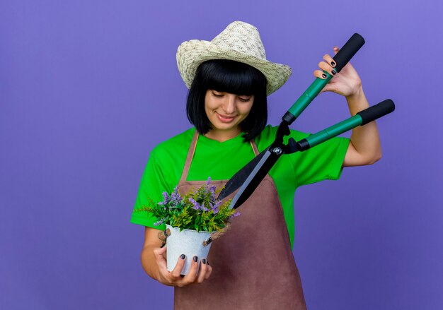 Lieta giovane giardiniere femminile in uniforme che indossa il cappello da giardinaggio tiene i clippers sui fiori in vaso di fiori