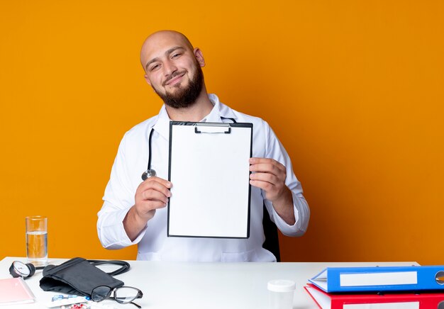 Lieta giovane maschio calvo medico indossando abito medico e uno stetoscopio seduto alla scrivania con strumenti medici tenendo appunti isolato su sfondo arancione