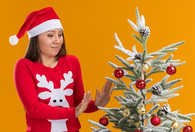 Lieta giovane ragazza asiatica che indossa il cappello di Natale con maglione in piedi vicino albero di Natale isolato su sfondo arancione
