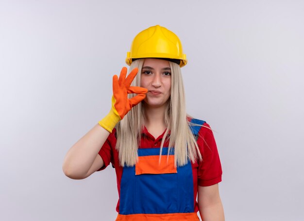 Lieta giovane ragazza bionda ingegnere costruttore in uniforme indossando guanti facendo segno ok su uno spazio bianco isolato con spazio di copia